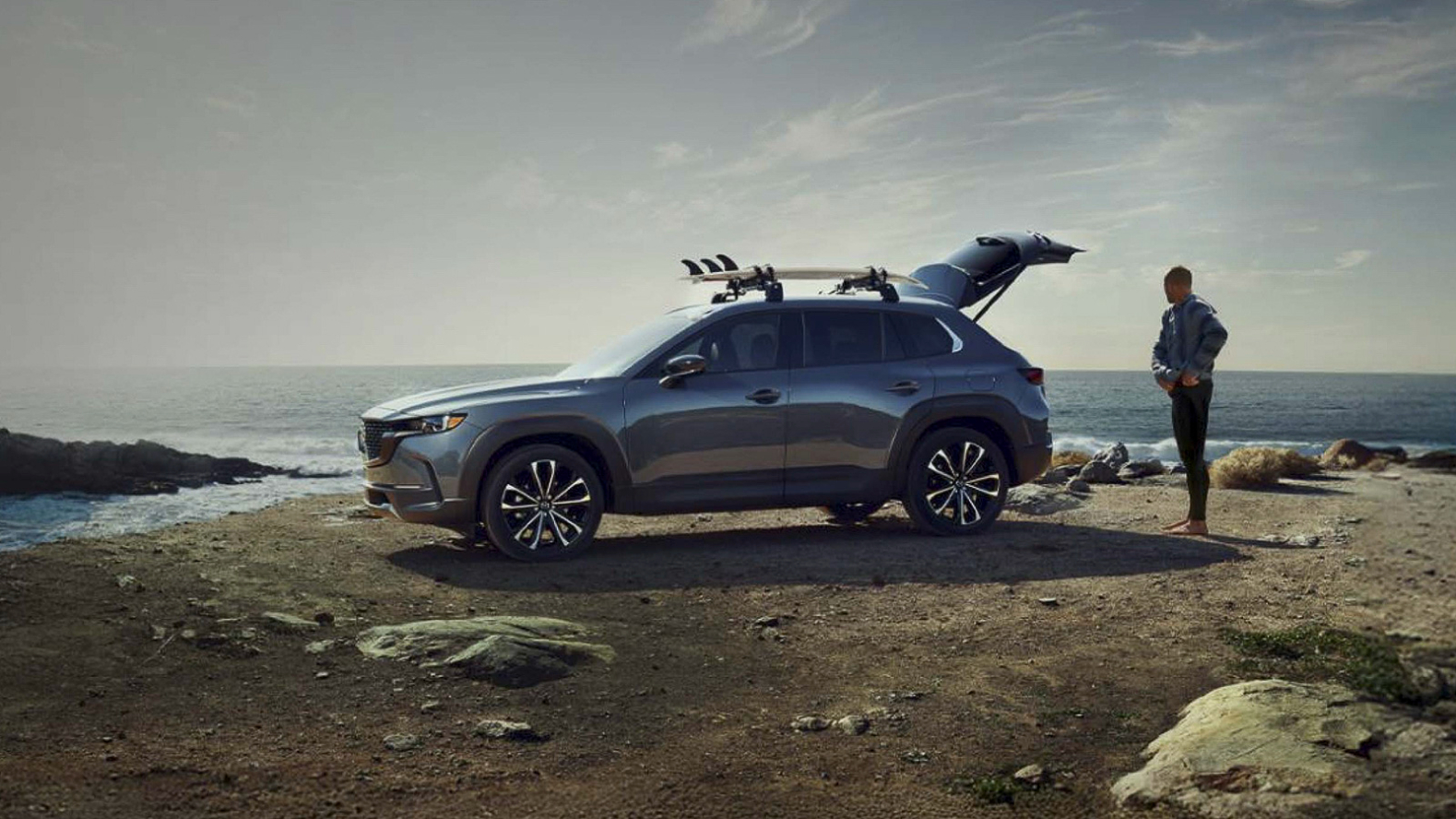 A silver Mazda CX-50 parked on cliffside near the ocean with the trunk open as a man stands behind it.