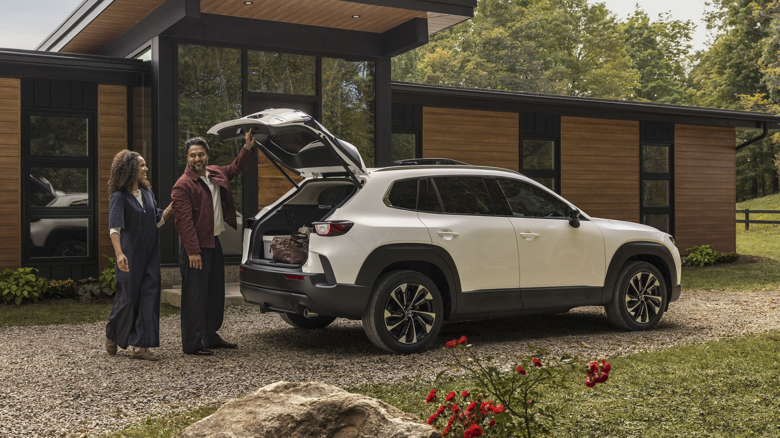 A couple loading up the back of their white Mazda CX-50 Hybrid, parked in front of a small house.
