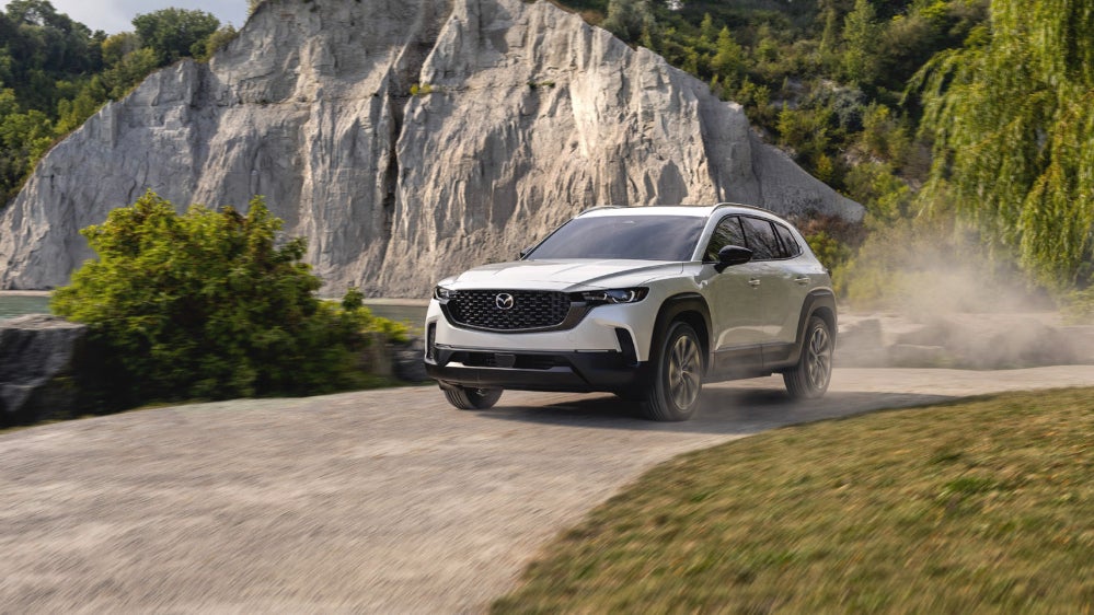 A silver Mazda CX-50 Hybrid driving down a dirt road near a mountain.
