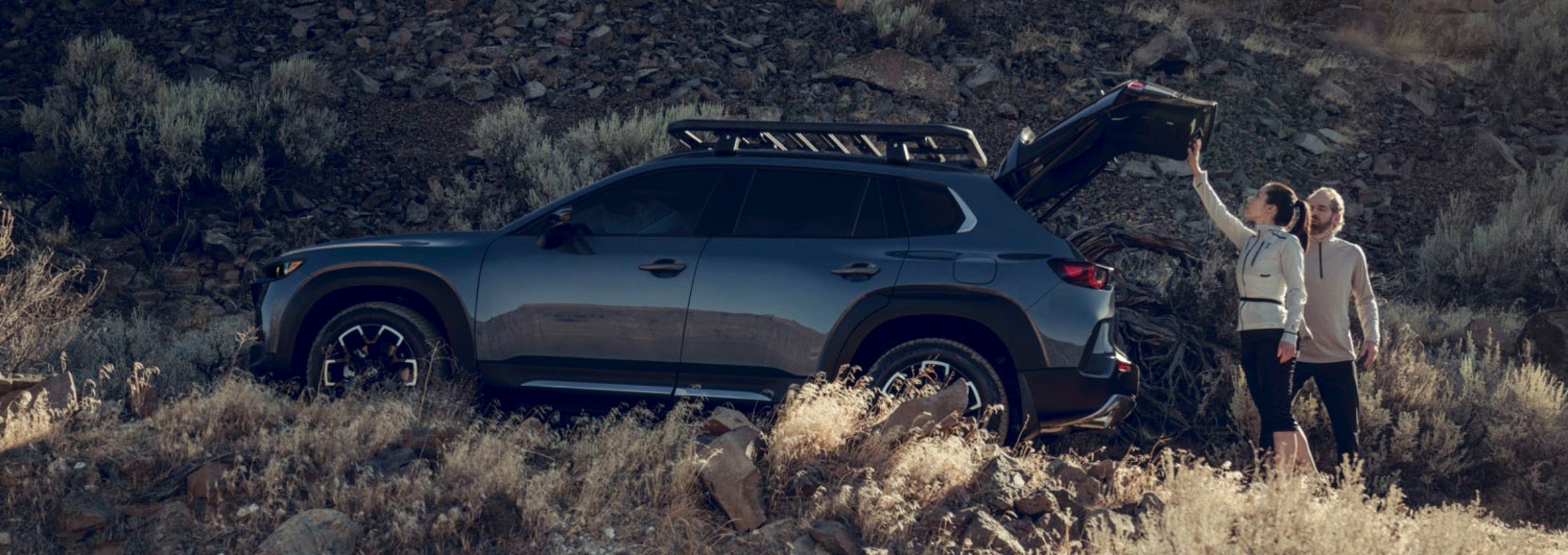 A couple checking the trunk of their Mazda CX-50, which is parked on hill in a desert-like area.