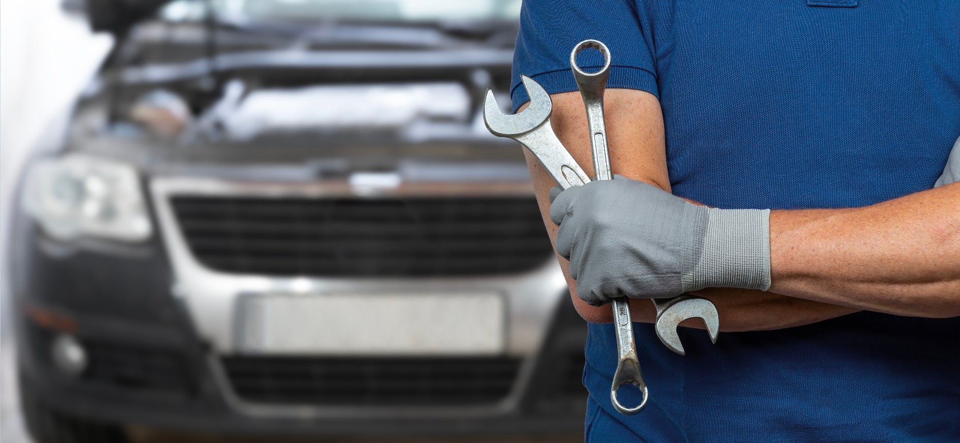 A technician with crossed arms holding wrenches in front of a gray vehicle.